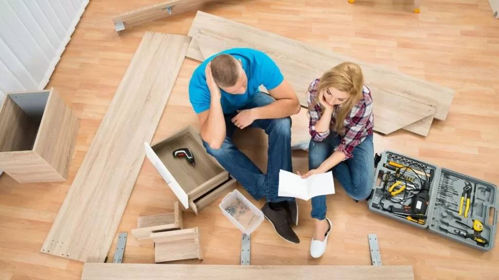 como guardar móveis desmontados para não empenar: casal sentado no chão com um monte de peça de moveis e eles lendo o manual.