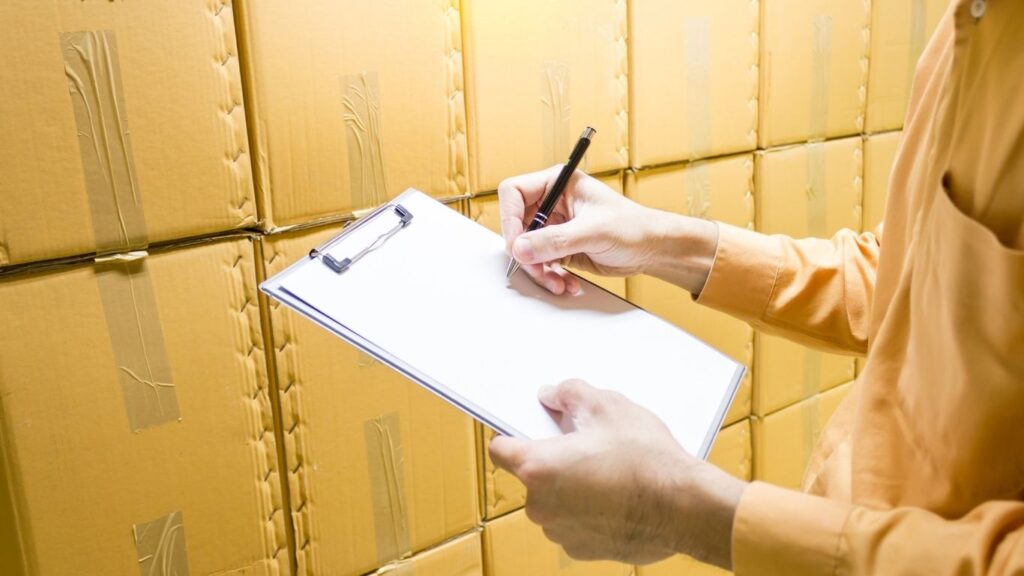 pessoa anotando algo em prancheta enquanto olha caixas